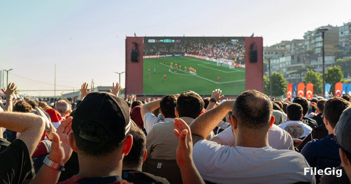 a packed football stadium under broadcast lights, seen from the upper tier, with camera rigs visible on the sidelines